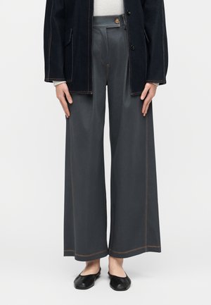 Person iført grå bukser med vide ben og synlige orange syninger, sorte flade sko, en sort jakke og en hvid sweater.