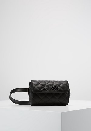 Sac banane en cuir matelassé noir avec sangle ajustable et logo "GUESS" sur le rabat avant, présenté sur une surface blanche avec un fond gris.