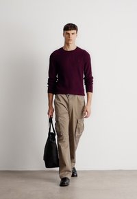 Mann trägt einen kastanienbraunen Strickpullover und beige Cargo-Hosen und hält eine schwarze Tragetasche. Der Boden ist neutral und der Hintergrund ist schlicht.