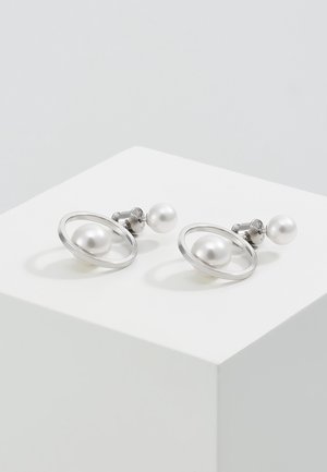 Boucles d'oreilles puces en argent avec un petit support en perle et une plus grande perle encadrée dans un cercle en argent sur une surface blanche.