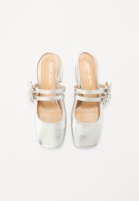 Scarpe slingback metalliche argento con punta quadrata, due cinturini e un accento di gioiello sul lato. Interno morbido e tacco modesto.