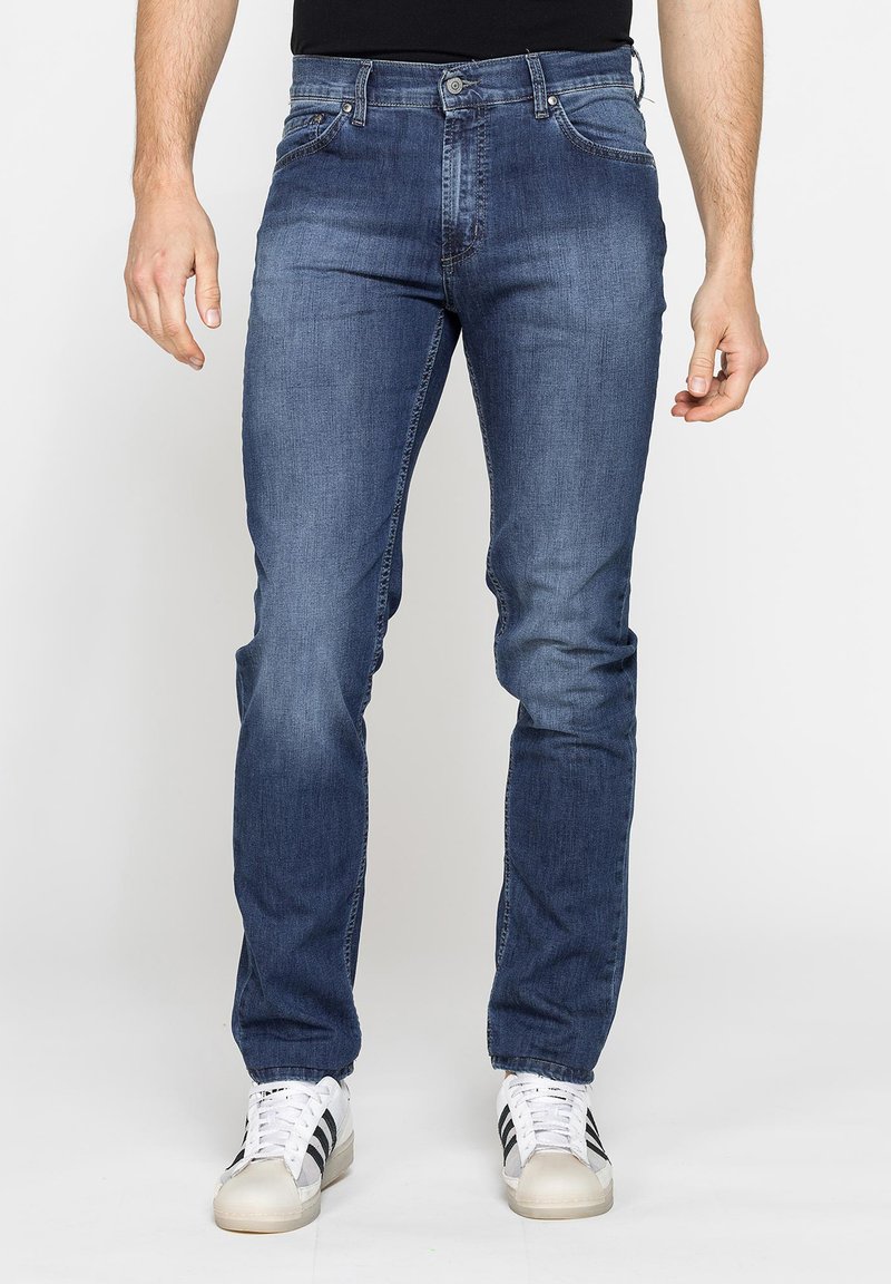 Uomo che indossa jeans slim-fit blu e sneakers bianche con strisce nere, in piedi davanti a uno sfondo chiaro uniforme.