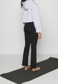 Haut à capuche lavande raccourci, associé à des leggings noirs évasés. Pieds nus sur un tapis de yoga noir texturé.