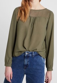 Blusa verde oliva con maniche lunghe e un sovrapposto in pizzo testurizzato sul collo, abbinata a pantaloni in denim blu a vita alta.
