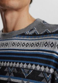 Jersey gris con patrones geométricos en azul, negro y gris. Cuello redondo, tejido texturizado y detalle sutil del logo en el pecho.