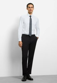 White, long-sleeve shirt; dark patterned tie; black trousers; glossy black dress shoes; figure standing with one hand in pocket against a plain background.