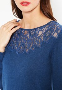 Sininen neulepusero, jossa on aaltoileva pitsikaulus ja pitkät hihat, sileä pinta ja herkullinen kukkakuosi.