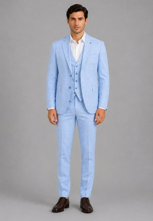 Homme debout portant un costume trois pièces bleu clair avec une chemise blanche et des chaussures habillées marron foncé sur un fond gris uni.