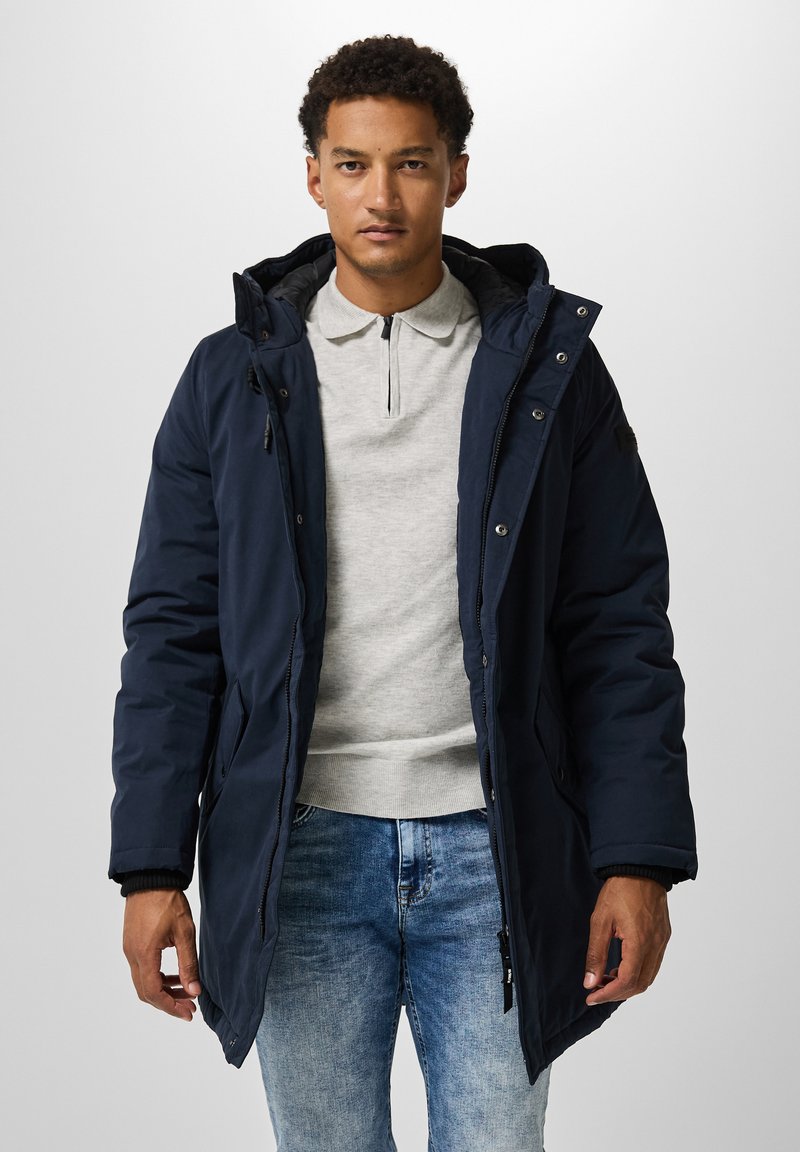 Parka acolchada en azul marino con capucha, cierre de cremallera y botones a presión. Camiseta polo gris debajo, combinada con jeans de un tono azul claro.