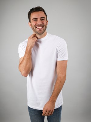 Lächelnder Mann in weißem T-Shirt und Jeans steht vor grauem Hintergrund, berührt den Hals mit der linken Hand und schaut leicht nach rechts.