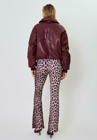 Giacca bomber in ecopelle color marsala con un logo in rilievo sulla schiena, abbinata a pantaloni a zampa d'elefante in stampa leopardata rosa. Texture liscia e design aderente.