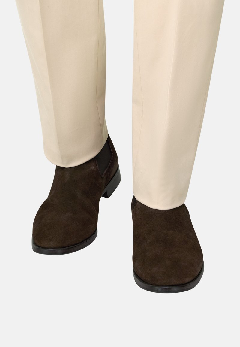 Bottes Chelsea en daim marron foncé portées avec un pantalon beige, montrant les vues de face et de côté des deux pieds.