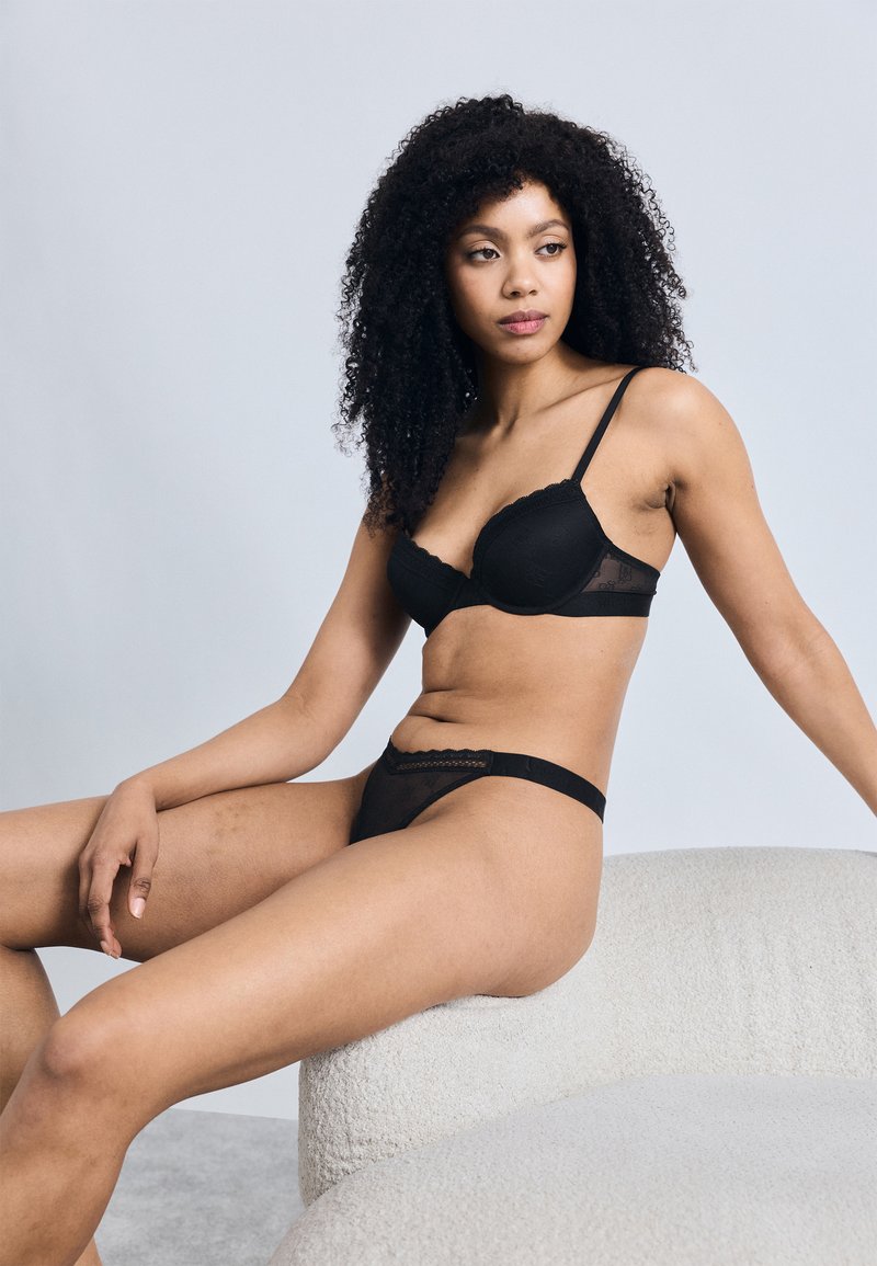 Σετ μαύρα εσώρουχα με ενισχυμένο σουτιέν διακοσμημένο με δαντέλα και αντίστοιχο διάφανο στρινγκ, τοποθετημένα πάνω σε μία ανοιχτού χρώματος υφή επιφάνειας.