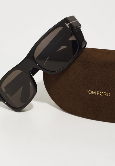 Occhiali da sole neri con montatura quadrata, lenti scure e dettagli hardware sottili, accanto a una custodia in camoscio marrone con la scritta "TOM FORD" impressa.