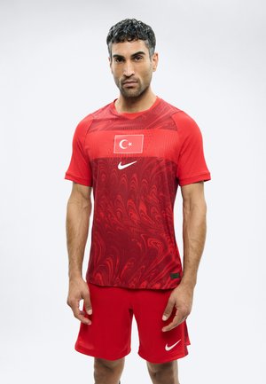 Άνδρας στέκεται φορώντας κόκκινα αθλητικά ρούχα της Nike με τη σημαία της Τουρκίας στο στήθος, που αποτελείται από κοντομάνικο μπλουζάκι και ταιριαστό σορτς, πάνω σε απλό φόντο.