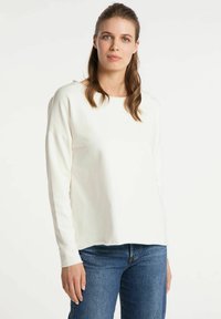 Haut crème à manches longues avec un col rond, épaules tombantes et coupe décontractée, associé à un jean bleu. Tissu doux avec une texture minimale.