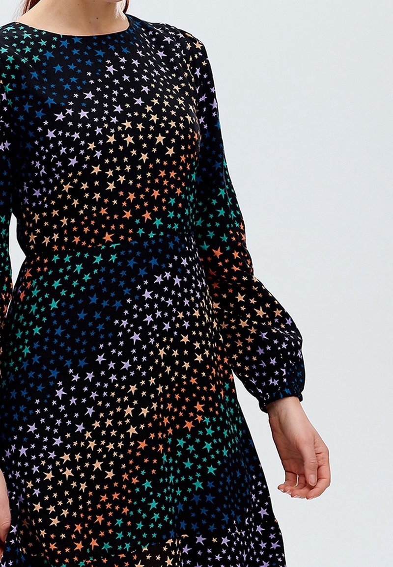 Schwarzes Kleid verziert mit mehrfarbigen Sternmustern, ausgestattet mit langen Ärmeln mit gerafften Manschetten und einem runden Ausschnitt.