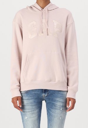 Persoon die een lichtroze GAP-hoodie met voorkantzak draagt en een blauw vervaagde gescheurde jeans, staand tegen een eenvoudige achtergrond.