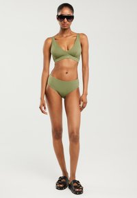 Oliwkowy strój kąpielowy z trójkątnym topem i wysokim stanem, w zestawie z czarnymi sandałami z złotymi akcentami.