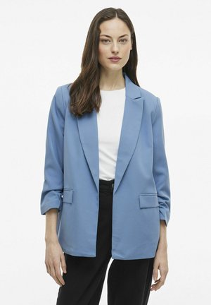 Světlomodrý na míru šitý blazer s klopovým límcem, zapínáním na jeden knoflík a dvěma předními kapsami. Otočené rukávy ukazují jemnou texturu látky.