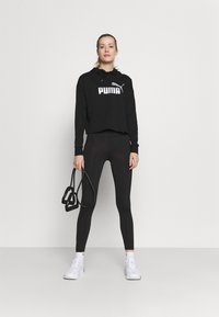 Sudadera corta negra con el logo "PUMA", combinada con leggins negros y zapatillas deportivas blancas; la modelo sostiene bandas de resistencia negras.