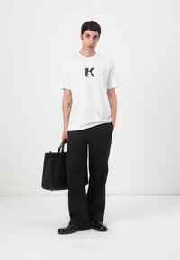 T-shirt branco de mangas curtas com gráfico preto "K", combinado com calças largas pretas e uma bolsa de pele preta. Texturas suaves em todo o conjunto.