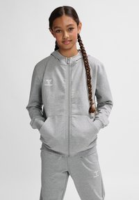 Grauer Kapuzenpullover mit Reißverschluss aus weichem Stoff, ausgestattet mit einer Kapuze, Fronttaschen und einem weißen Logo auf der linken Brustseite. Kombinierbar mit passender Jogginghose.