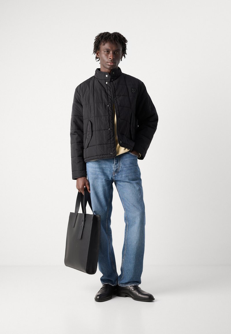 Veste matelassée noire avec un col haut, portée sur un haut jaune clair. Jean en denim bleu et chaussures en cuir noires. Tient un sac fourre-tout noir.