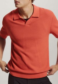 Jeune personne portant un polo rouge-orange texturé à manches courtes avec col en V et pantalon noir, mains près de la taille.