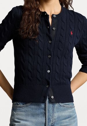 Femme portant un cardigan bleu marine en maille torsadée avec des boutons et un logo rouge sur la poitrine, associé à un jean bleu clair, mains sur les hanches.
