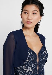 Giacca bolero in chiffon blu navy sopra un abito in pizzo dettagliato con complessi ricami in perline e scollatura profonda.