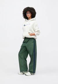 Camisola branca com capuz e logo azul-marinho, combinada com calças largas verdes com uma risca azul-marinho. Tênis brancos e verdes completam o visual.