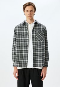 Tjekket flannelsskjorte i sort, grå og hvid med knaplukning foran og en brystlomme med skråternet mønster.