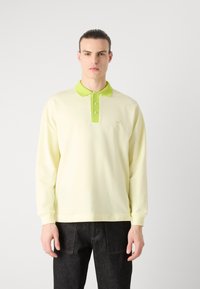 Camisa polo de manga larga en amarillo claro con un cuello verde brillante, tapeta de tres botones y un sutil logo bordado en el lado izquierdo del pecho.