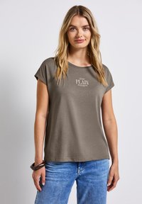 T-shirt en coton beige avec une coupe décontractée, des manches courtes retroussées et un logo imprimé "Assez SIMPLE" sur le devant. Détails de tissu texturé visibles.