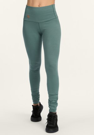 Legging in gedempt teal met hoge tailleband en subtiele logo, gecombineerd met zwarte sportschoenen op een neutrale achtergrond.