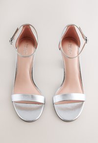 Sandales argentées ouvertes à l'avant avec brides de cheville et boucles, semelles plates et semelles intérieures beiges, placées côte à côte sur un fond clair.