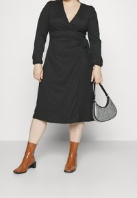 Vestido negro de manga larga con escote en V, atado a la cintura. Combinado con botas de tobillo brillantes de color marrón y un bolso negro con tachuelas.