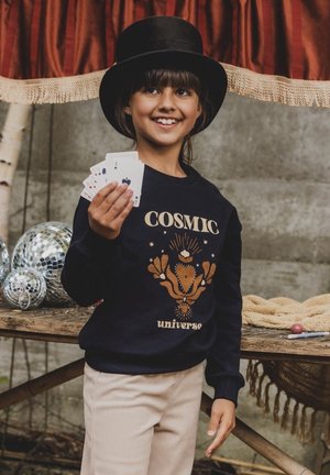 Marine sweatshirt met een 'COSMIC universe' graphic in goud, gecombineerd met lichtbeige broek. Kind houdt speelkaarten vast en draagt een zwarte hoge hoed.