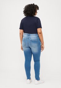 Person with curly hair facing away, wearing a navy t-shirt and light blue jeans, standing against a plain white background.