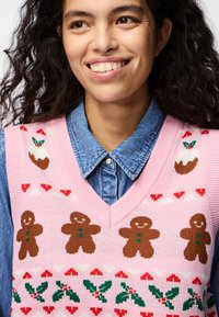 Chaleco de punto rosa con figuras de pan de jengibre, acebo y patrones de corazones. Se lleva sobre una camisa de mezclilla con cuellos.