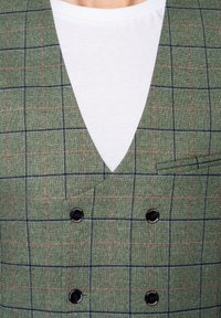 Zielona kamizelka w kratę z głębokim dekoltem w serek, podwójnym zapięciem na guziki, czarnymi guzikami i subtelną teksturą. Noszona na białej koszuli.