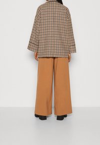Giacca oversize a scacchi marroni con maniche ampie, abbinata a pantaloni beige a gamba larga. Footwear neri visibili.