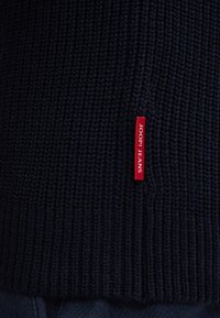Marinefarbener Strickpullover mit geribbelter Textur und einem auffälligen roten Etikett mit der Aufschrift "JOOP! JEANS" am Saum.