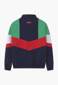 Marinblå og grøn vindjakke med rød og hvid chevronmønster. Har en høj krave og elastiske manchetter. Logo på ryggen.