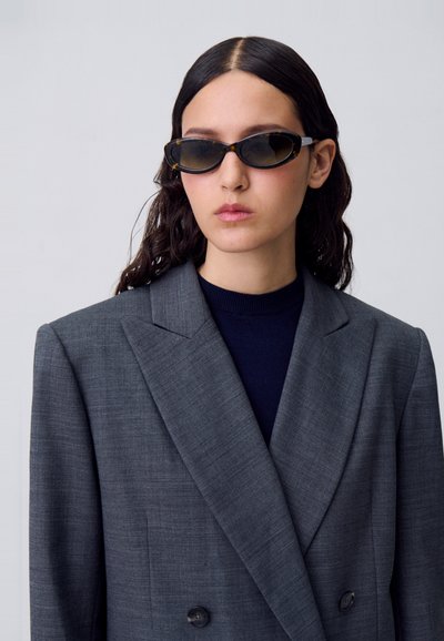 Blazer grigio doppiopetto con ampi revers, tessuto strutturato e due bottoni, abbinato a occhiali da sole neri ovali e a un dolcevita blu navy.