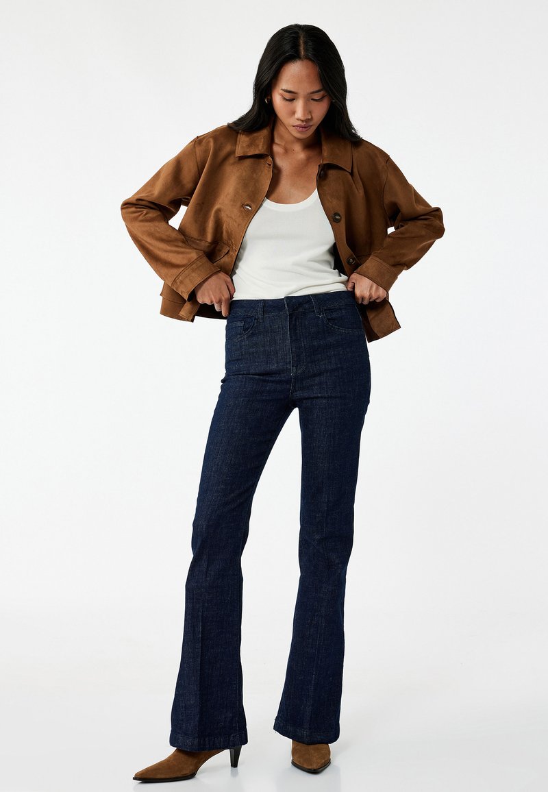 Bruine suède jas, witte tanktop, hoog in de taille gesneden donkere denim flare jeans, bruine haklaarzen, minimale knopen op de jas.