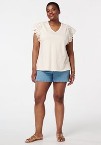 Haut beige à col en V avec des empiècements en crochet sur les manches volantées, associé à un short en denim bleu. Sandales à lanières marron clair. Textures lisses.