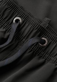 Zwarte zwembroek met een trekkoordontwerp, elastische tailleband en metalen oogjes met merkdetaillering. Zachte, gladde stoftextuur.
