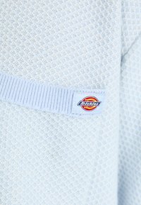 Šviesiai mėlynas tekstūruotas megztinis su juostu apačia ir žyma su Dickies logotipu apvalioje formoje.